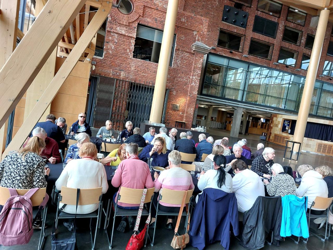 Kuva Sibeliustalon Metsähallista. Koko ryhmä kahden pitkän pöydän ääressä syömässä. Taustalla kaksi puista pylvästä korkeassa hallissa, sekä punaisen tiiliseinä, joka erottaa metsähallin lounasravintolasta.